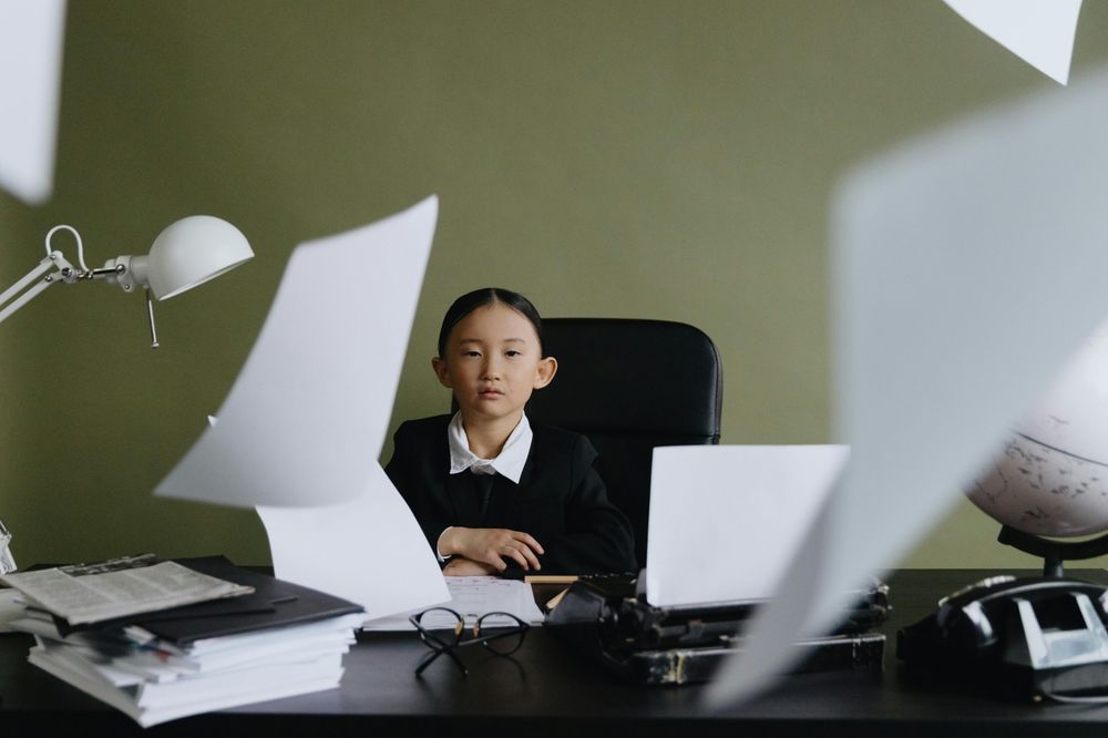 A young girl in a blazer sitting at a desk while papers fly through the air
