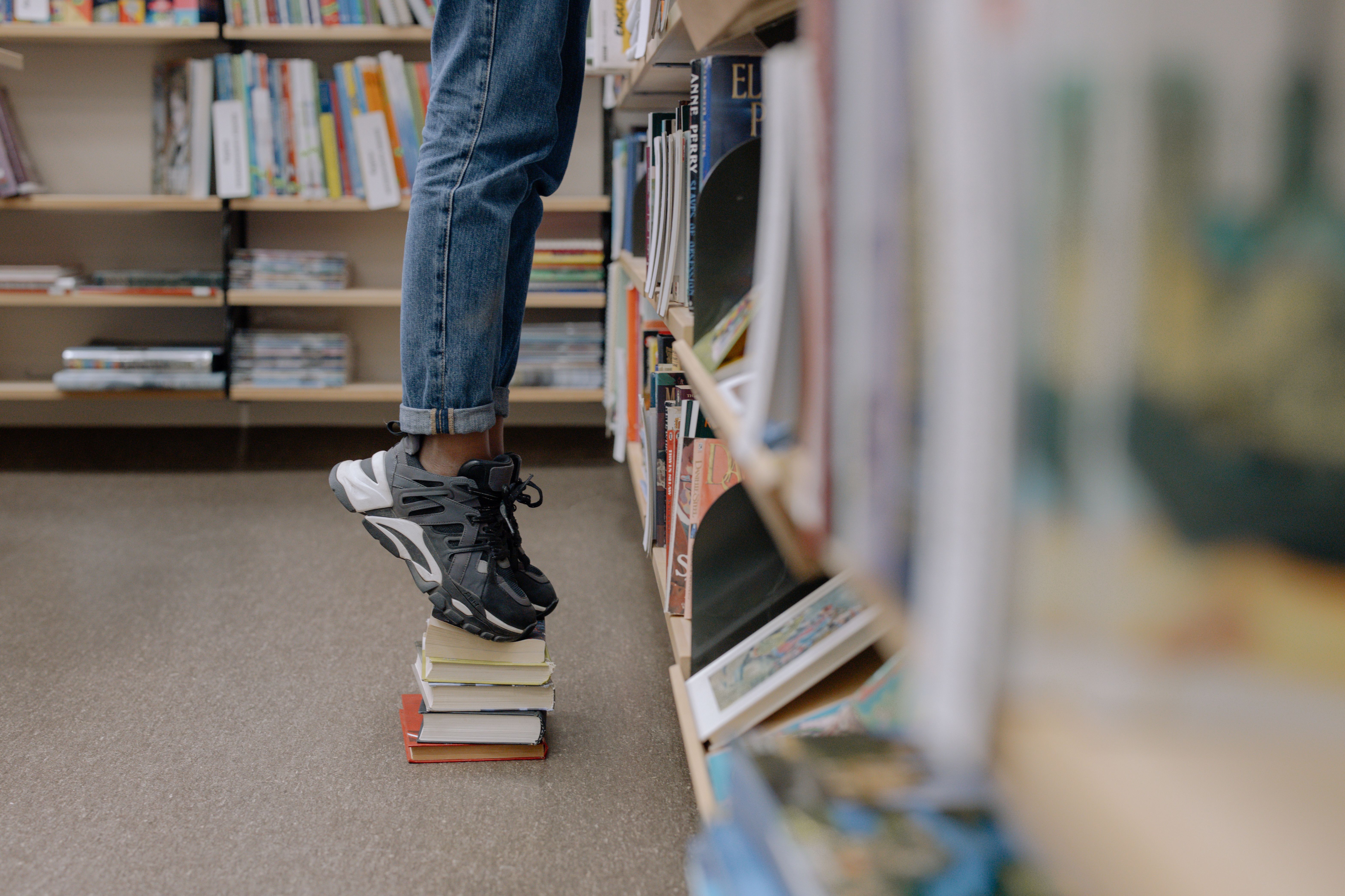 A child stands on a stack of books to reach a high library shelf.